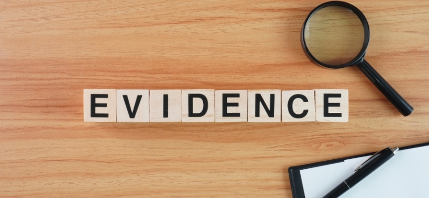 Wooden blocks spelling the word "EVIDENCE" next to a magnifying glass and a clipboard on a wooden surface, illustrating the investigative requirements for a sexual assault case.