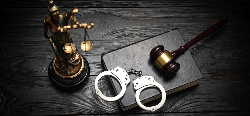 A metal pair of handcuffs, a judge's wooden gavel, and a law book placed next to a Lady Justice statue, representing charges of murder attempted under Canadian criminal law.
